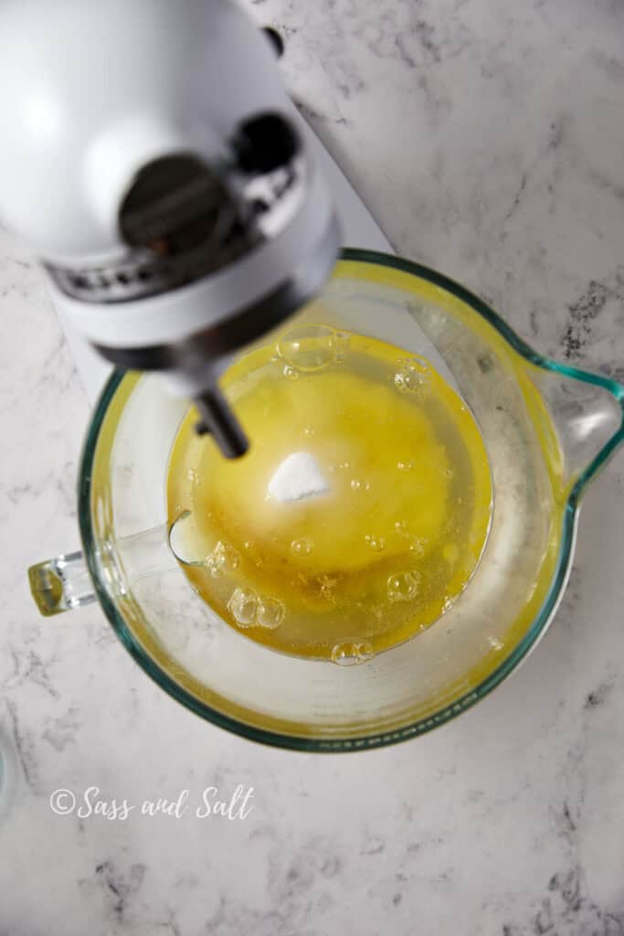 Top-down view of a stand mixer with a glass mixing bowl containing a blend of egg whites, sugar and vanilla ingredients on a white marble countertop. "Sass and Salt" is written in the lower left corner.