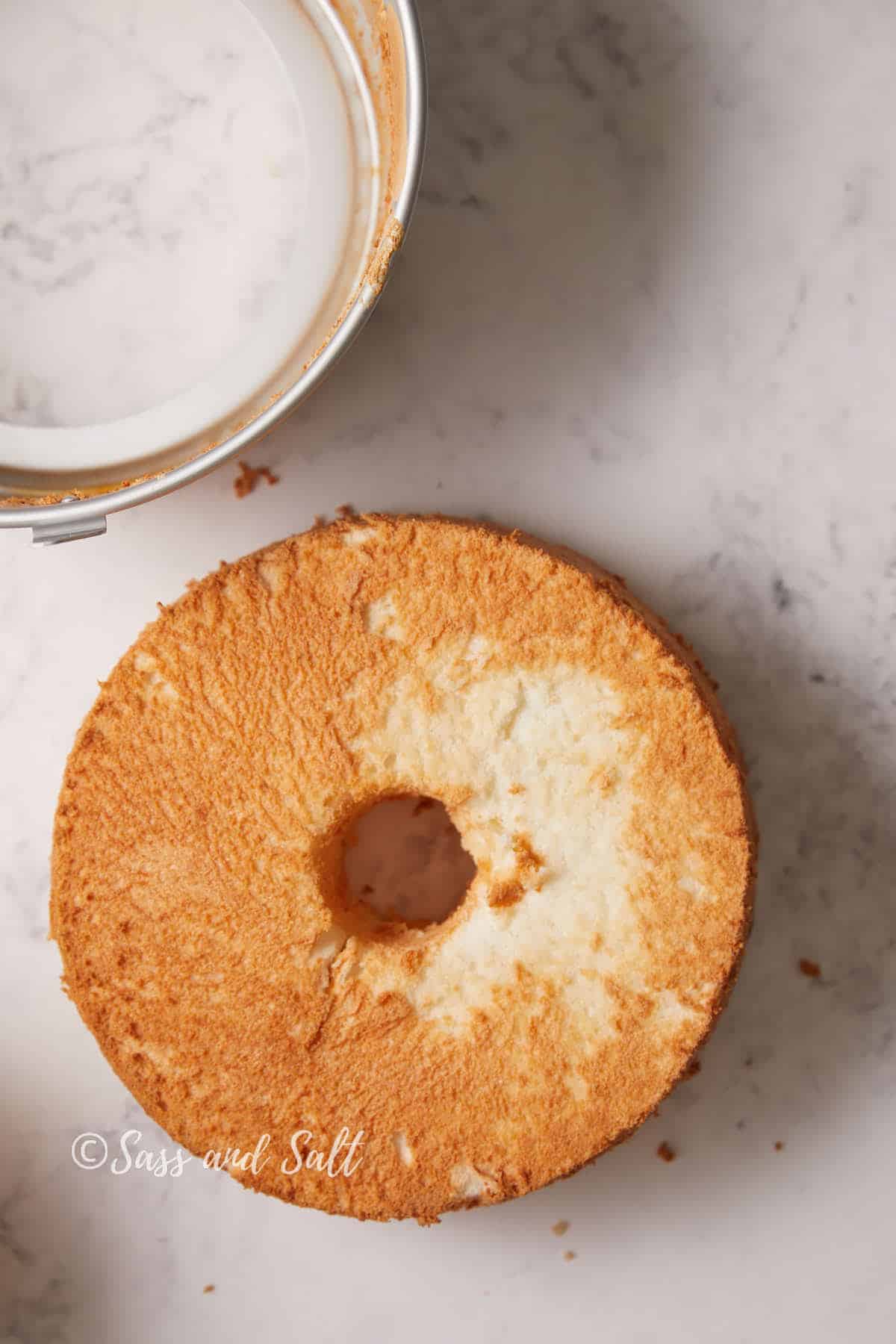 A golden brown angel food cake with a hole in the center sits on a white marble countertop. Part of a cake pan is visible at the top edge of the image. The cake looks fluffy and light with a slightly uneven surface. Some crumbs are scattered around.