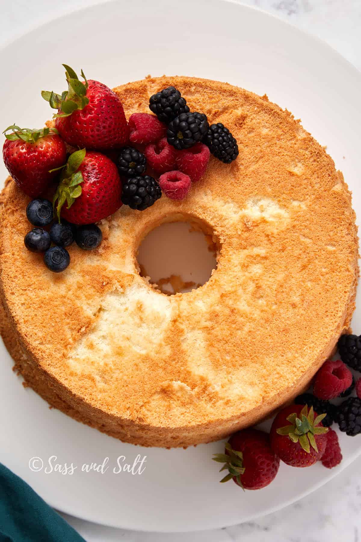 An angel food cake topped with an assortment of fresh berries, including strawberries, blueberries, raspberries, and blackberries. The cake is centered on a white plate, and a green cloth is partially visible at the edge.