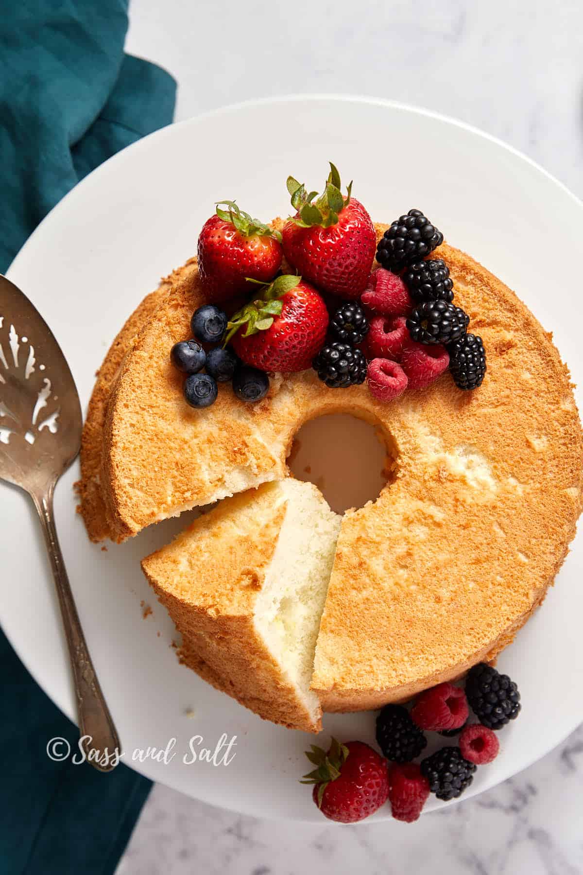 A round angel food cake with a slice missing, topped with fresh strawberries, blueberries, blackberries, and raspberries on a white plate. A serving utensil rests on the plate to the left. A piece of fabric corner is visible in the upper left corner.