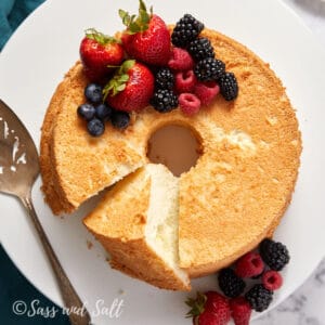 A ring-shaped, angel food cake with a slice cut out is displayed on a white plate. The cake is garnished with fresh strawberries, blueberries, raspberries, and blackberries. A metal cake server rests on the plate next to the cake. The image is signed "Sass and Salt.