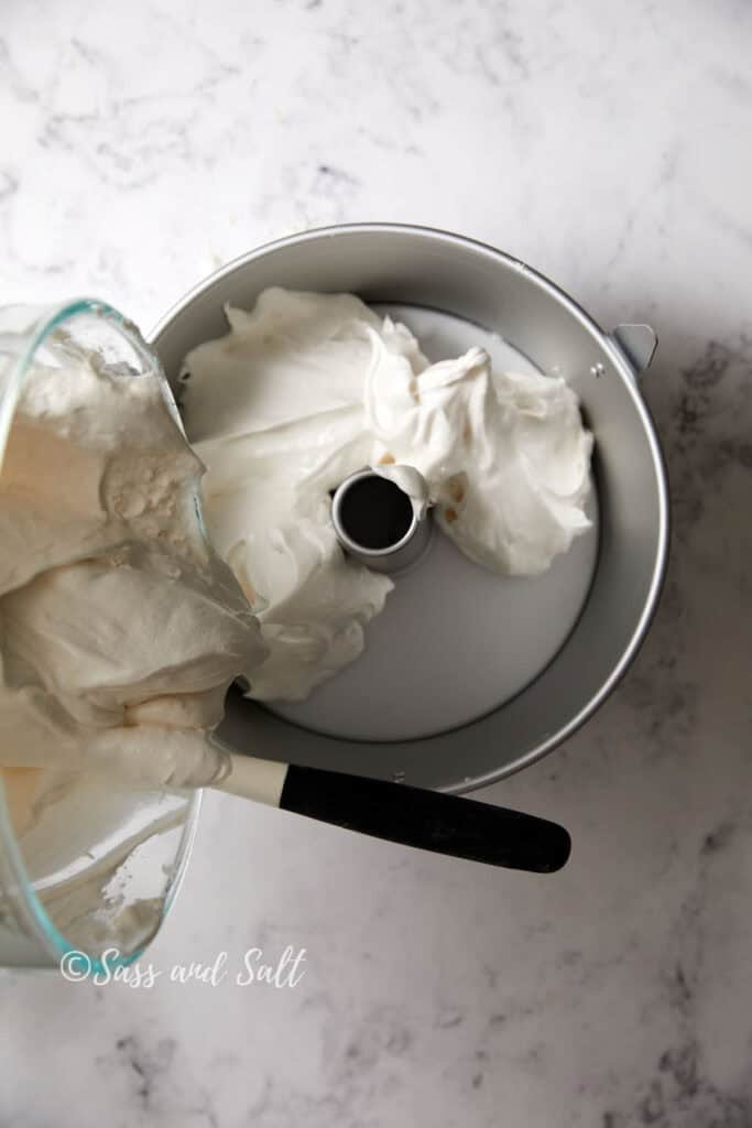 A glass bowl is pouring a thick, white batter into a metal tube pan on a marble countertop. A black-handled spatula is being used to scrape the mixture from the bowl into the pan. The image has a watermark reading "Suss and Salt.