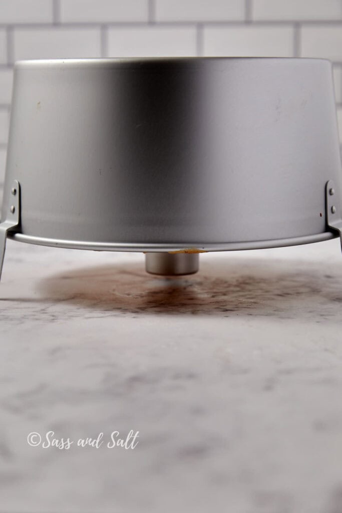 A silver bundt cake pan is upside down on a kitchen countertop. The countertop has a marble-like design, and the background features white subway tiles. There's a faint shadow and slight discoloration below the center of the pan.