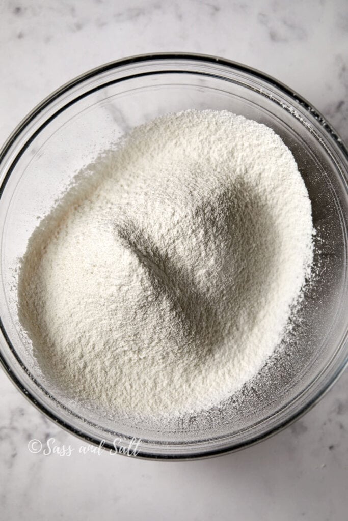 A clear glass bowl filled with a mound of fine, white, sifted flour sits on a marble countertop. The texture of the flour appears soft and powdery. The photo is watermarked with the text "Sass and Sill" in the bottom left corner.