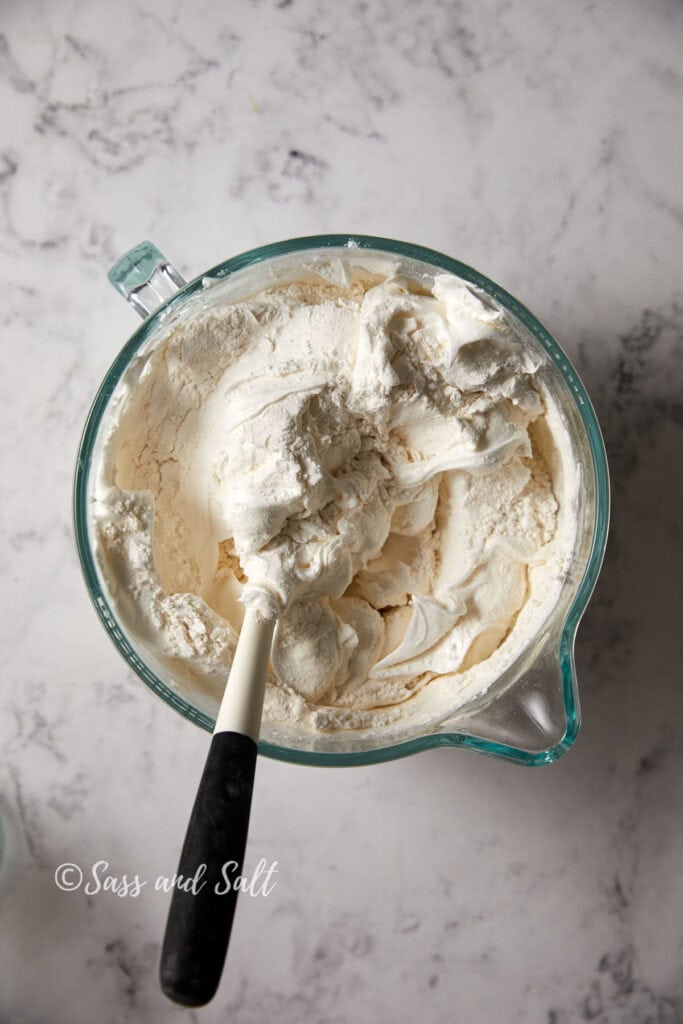 A glass mixing bowl filled the angel food batter and cake flour being mixed together with a black and white spatula. The bowl is placed on a light marble countertop. The image is signed "Sass and Salt" in the bottom left corner.