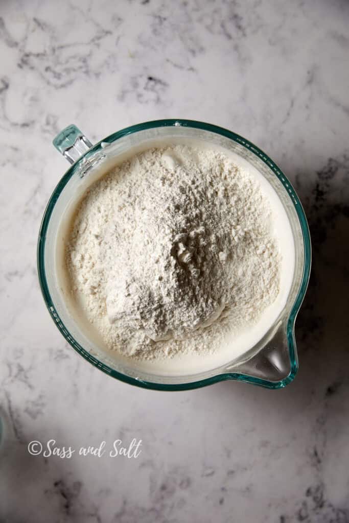 A top-down view of a glass mixing bowl on a marbled countertop, with cake flour on top of the egg white angel food batter.