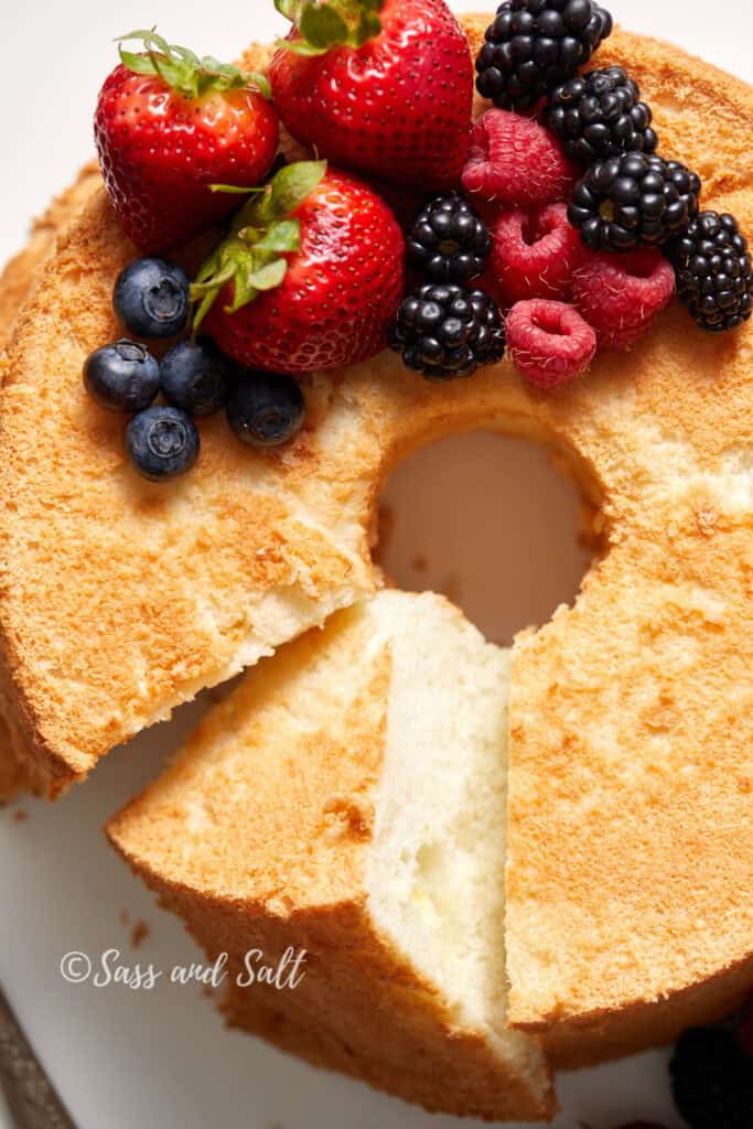 A fluffy, golden chiffon cake with a slice removed. The cake is adorned with a variety of fresh berries, including strawberries, raspberries, blackberries, and blueberries. The background is plain, highlighting the cake's airy texture and vibrant fruit topping.