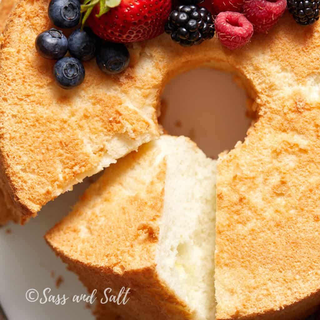 A fluffy angel food cake with a slice removed is topped with fresh berries, including strawberries, blueberries, raspberries, and blackberries. 