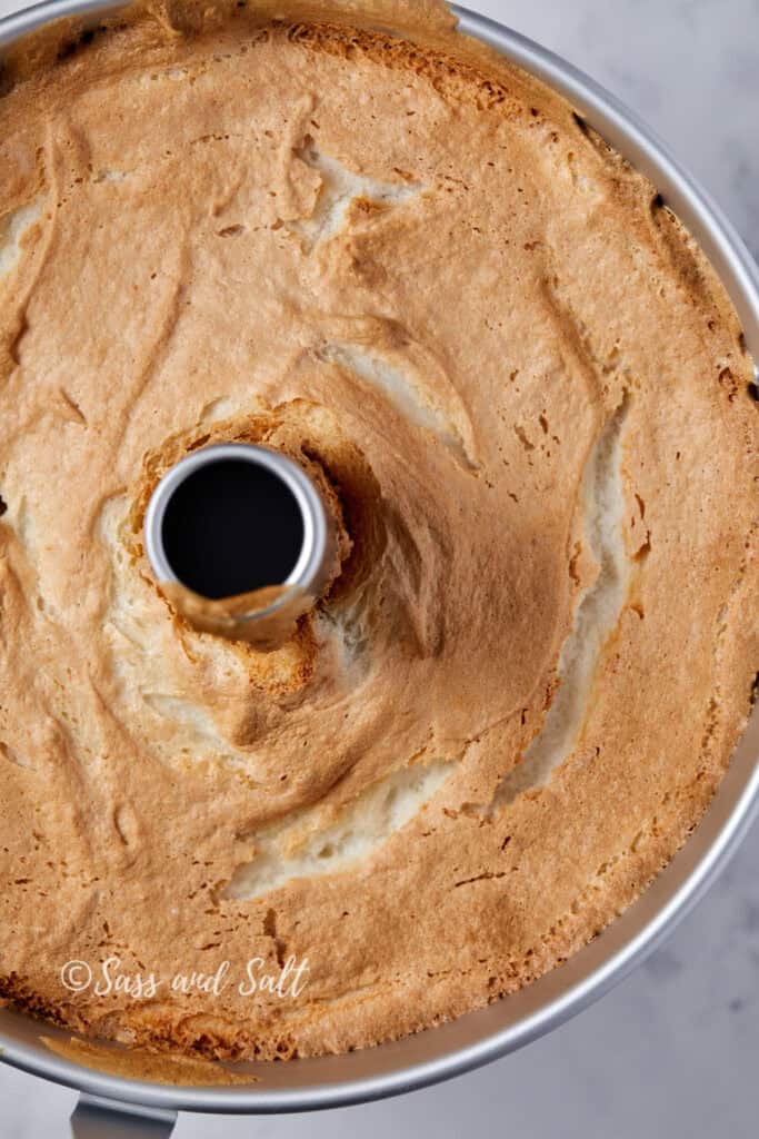 Close-up of a freshly baked angel food cake in a tube pan. The cake has a light golden-brown crust and a fluffy, airy texture. Visible cracks and crevices add to its homemade appearance. The photo is taken from above on a marble surface. The words "Sass and Salt" are at the bottom left corner.