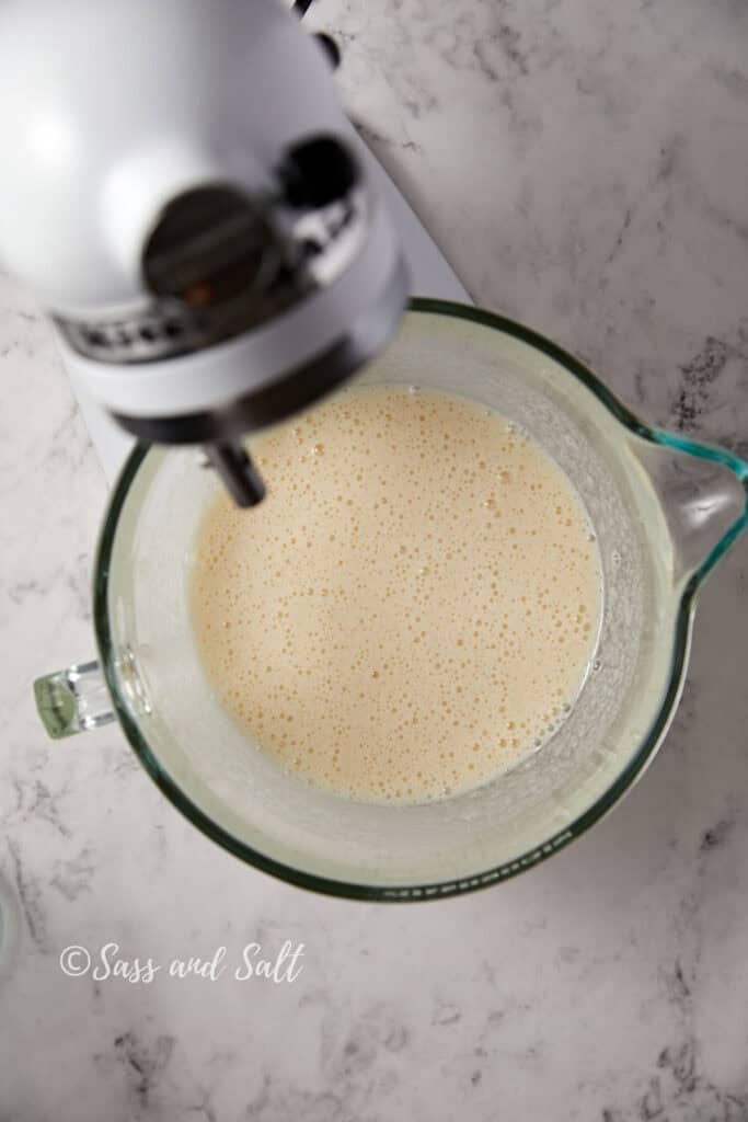 An overhead view of a mixer blending batter in a glass bowl. The mixer is white and the glass bowl shows a creamy mixture. The background is a marble countertop. The words "Sass and Salt" are at the bottom left corner of the image.