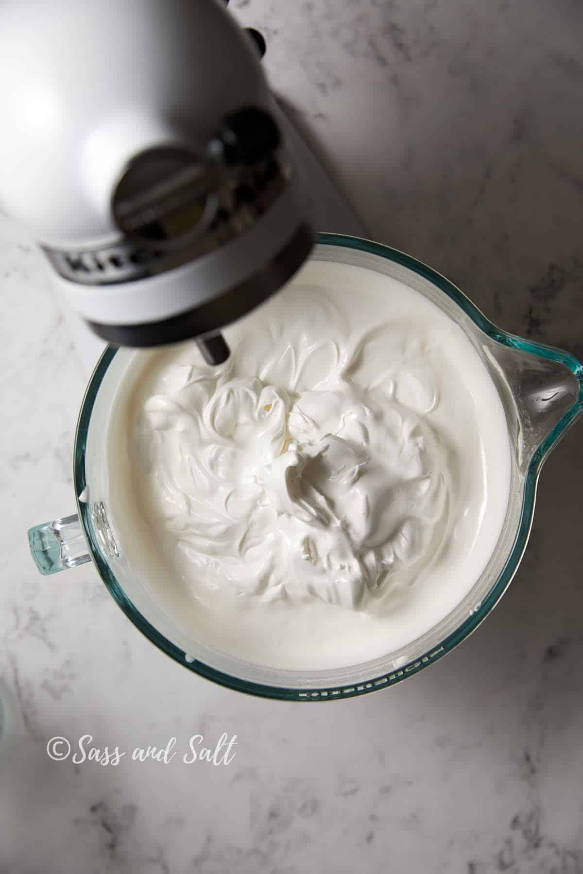 A glass bowl with freshly whipped egg whites, sugar, vanilla, and lemon juice sits under a white stand mixer. The creamy mixture appears fluffy and smooth. The scene is set on a marble countertop. There is a faint watermark that reads "Sass and Salt" on the left side.