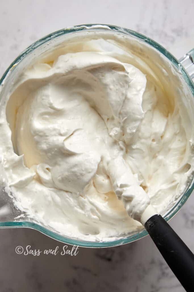 A glass bowl filled with creamy, whipped white frosting. A black and white spatula is partially submerged in the frosting, its handle resting on the bowl's edge. The background features a light, slightly marbled surface. "Sass and Salt" is written at the bottom of the image.