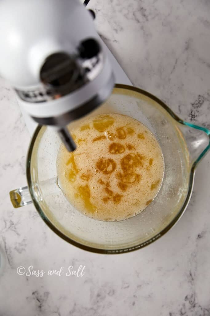 A top view of a stand mixer on a marble countertop. The mixing bowl contains the egg whites, sugar and vanilla after mixing for one minute. The image is watermarked with "Sass and Salt" in the lower left corner.