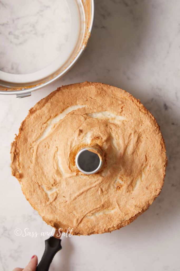 A top view of a freshly baked, golden-brown angel food cake in a tube pan on a marble countertop. The cake has been removed from the outer pan. The cake has a textured, fluffy surface. A hand is holding an offset spatula to loosen the bottom of the pan from the cake.  separated bottom part of the tube pan. 