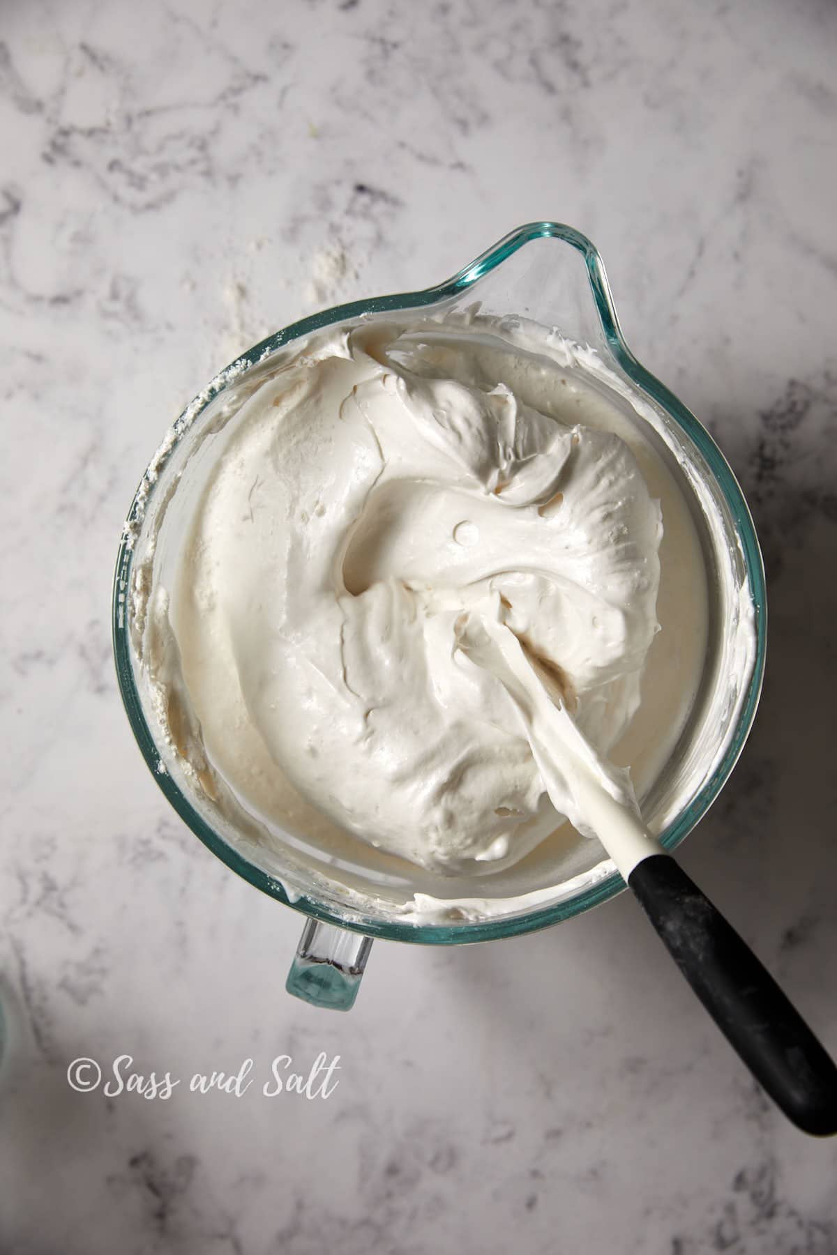 A glass bowl filled with fluffy, angel food cake batter sitting on marble countertop. A rubber spatula rests in the bowl, partially covered in the batter. The logo "Sass and Salt" is visible in the lower left corner.