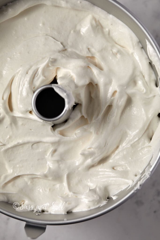 A close-up image of the angel food cake batter in a tube pan. The creamy batter has a light, smooth texture and is evenly spread. The center tube of the pan is visible, surrounded by the batter. The pan appears to be ready for baking on a marble surface.