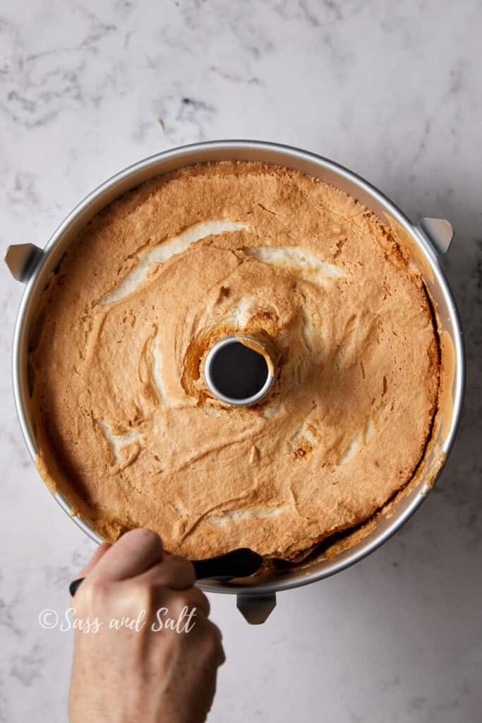 A person's hand holds an offset spatula running it around the edge of a freshly baked, angel food cake in a metal pan. The cake has a light brown, fluffy texture with a golden top. The background is a white, marble surface. The words "Sass and Salt" are visible in the bottom left corner.