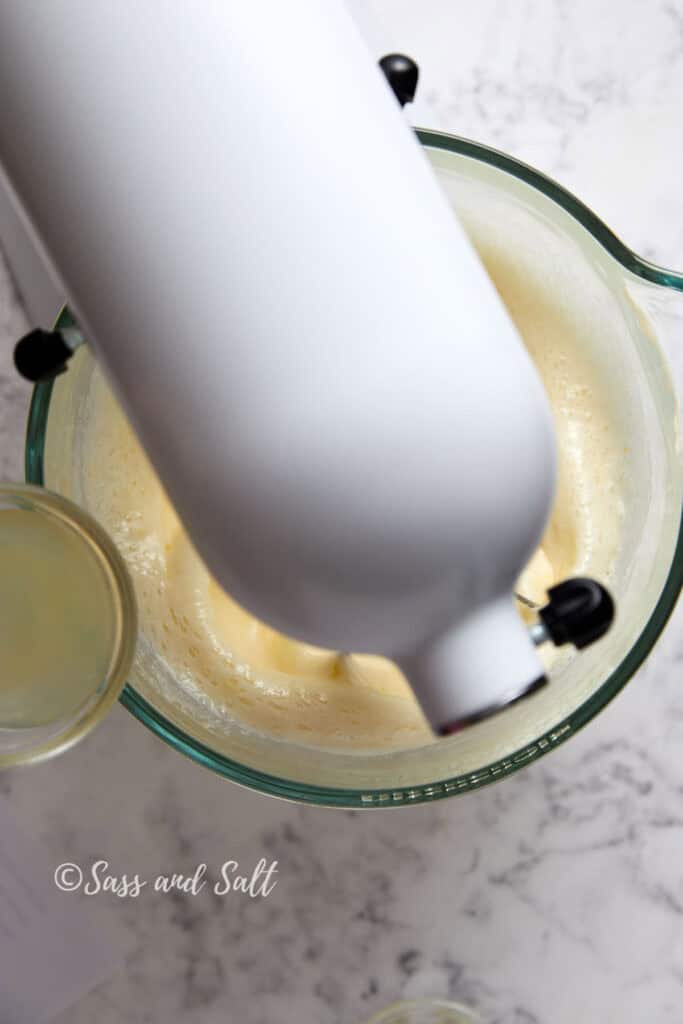 Top-down view of a stand mixer blending a creamy mixture in a glass bowl on a marble countertop. The mixer is white and the bowl contains a light yellow batter. A glass with lemon juice is partially visible beside the mixer. The text "Sass and Salt" is on the image.