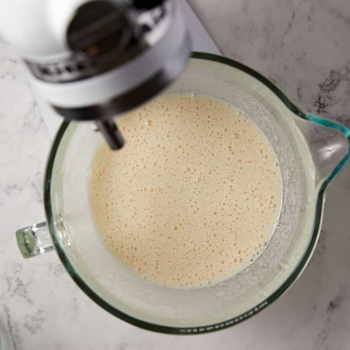 A glass mixing bowl filled with a light, frothy liquid mixture sits on a marble countertop next to the head of a stand mixer.