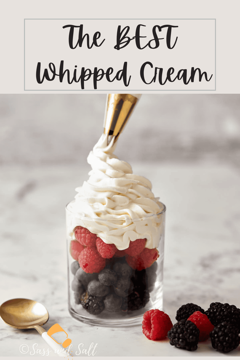 A glass filled with fresh raspberries, blueberries, and blackberries is being topped with a generous swirl of homemade whipped cream from a piping bag. A gold spoon rests beside the glass. The text on the image reads, "The BEST Whipped Cream.