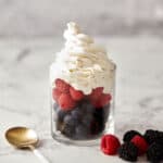 A glass filled with layered blueberries, raspberries, and blackberries, topped with a generous swirl of whipped cream. Beside the glass sits a gold and white spoon, with scattered blackberries and a raspberry on a white marble surface.