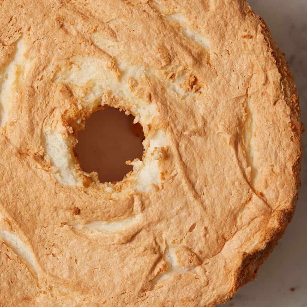 A close-up of a golden-brown angel food cake with a central hole and a light, fluffy texture on a marble surface.