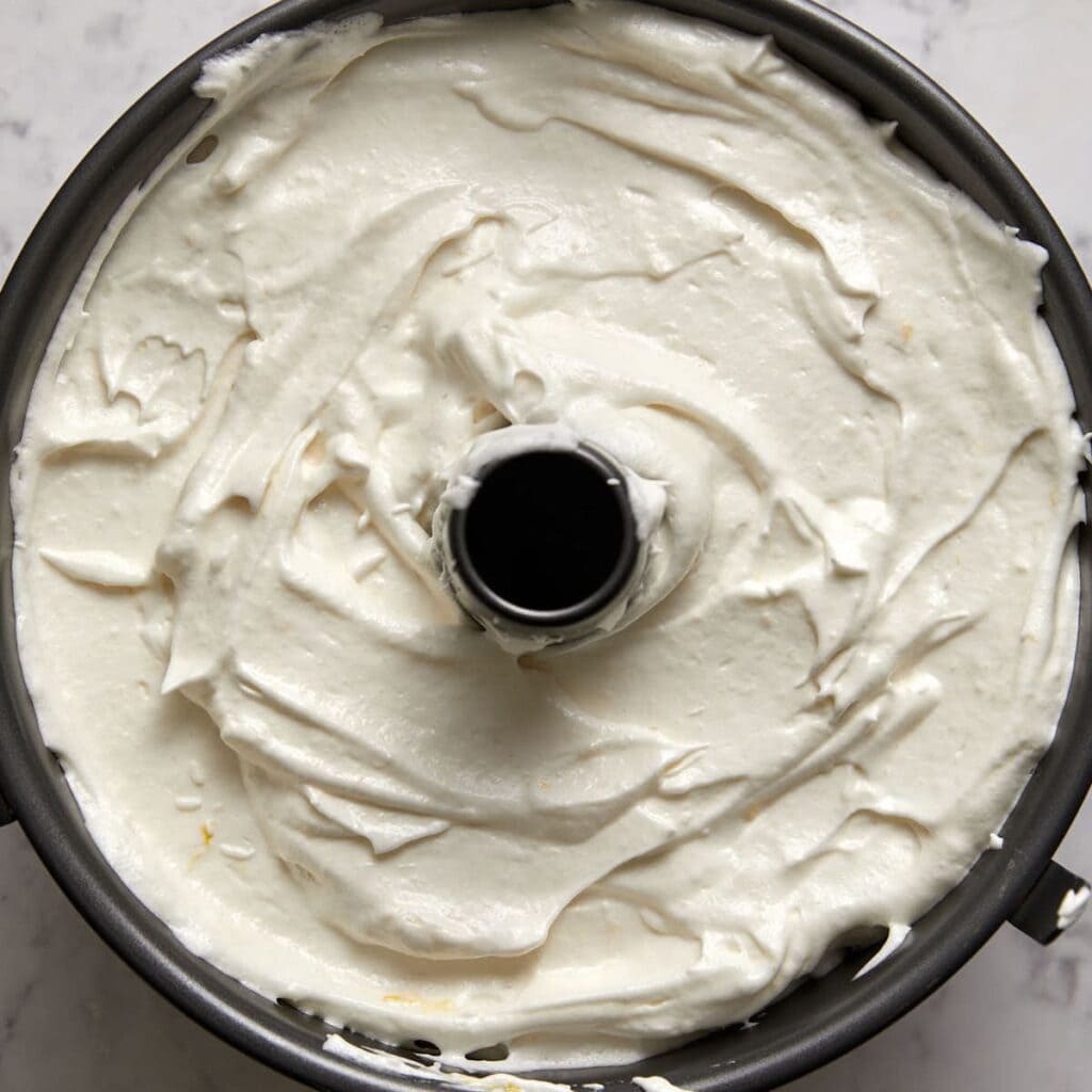 A close-up of a tube pan filled with smooth, fluffy angel food cake batter, spread evenly and ready to be baked, sitting on a marble surface.