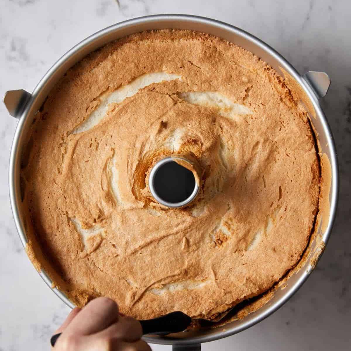 A hand runs a knife along the edge of a golden-brown angel food cake still inside a round tube pan on a marble surface, loosening the angel food cake.