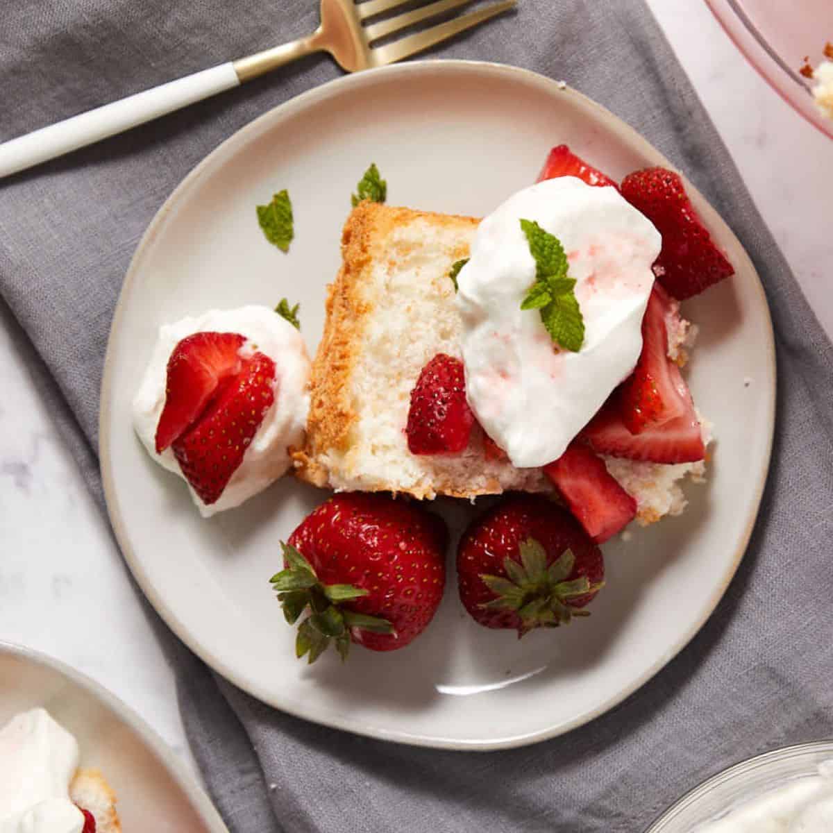 A slice of angel food cake with strawberries, topped with whipped cream and mint leaves, sits on a white plate. Whole strawberries and extra whipped cream are on the side, with a gold fork and gray napkin nearby.