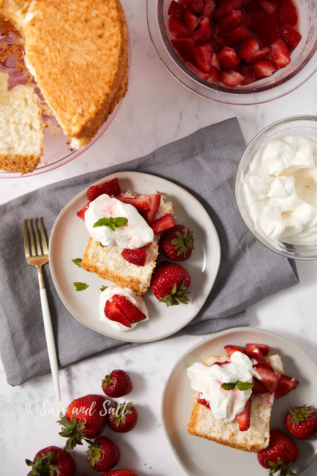 A flat lay of a plated strawberry shortcake dessert on a white plate with a fork on the side. The cake is topped with fresh strawberries and whipped cream. Additional strawberries, whipped cream, and a partially cut cake are nearby. The background is a gray cloth on a marble surface.