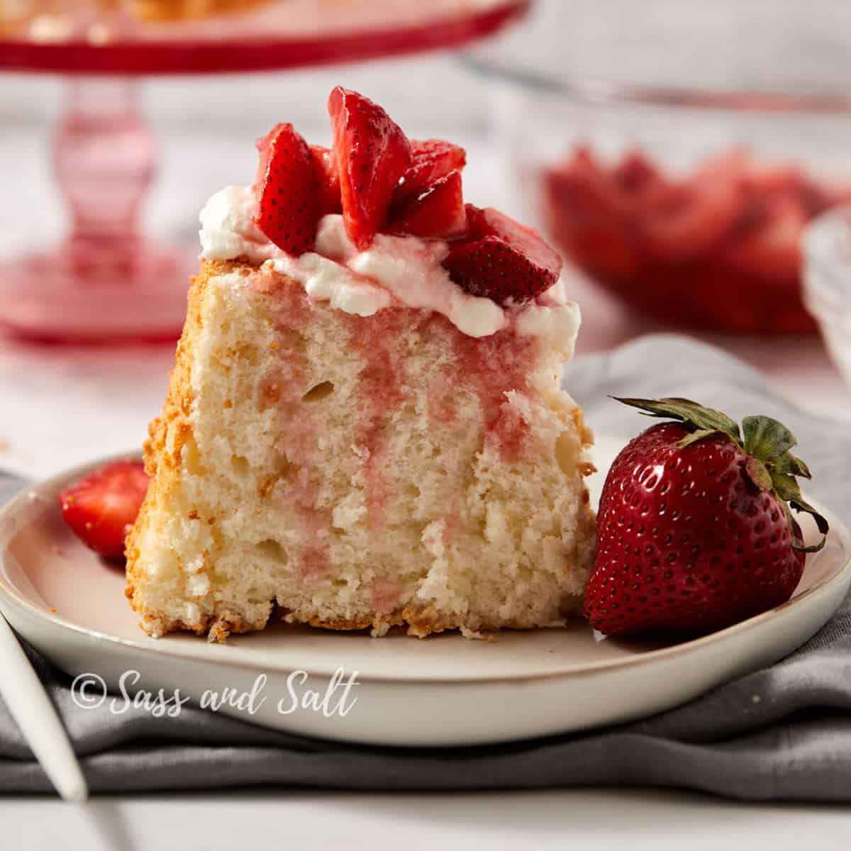 A slice of light, airy cake topped with whipped cream and fresh strawberry slices is served on a white plate. A whole strawberry is positioned beside the cake slice. The background includes a bowl of sliced strawberries and a red cake stand. (Credit: Sass and Salt).