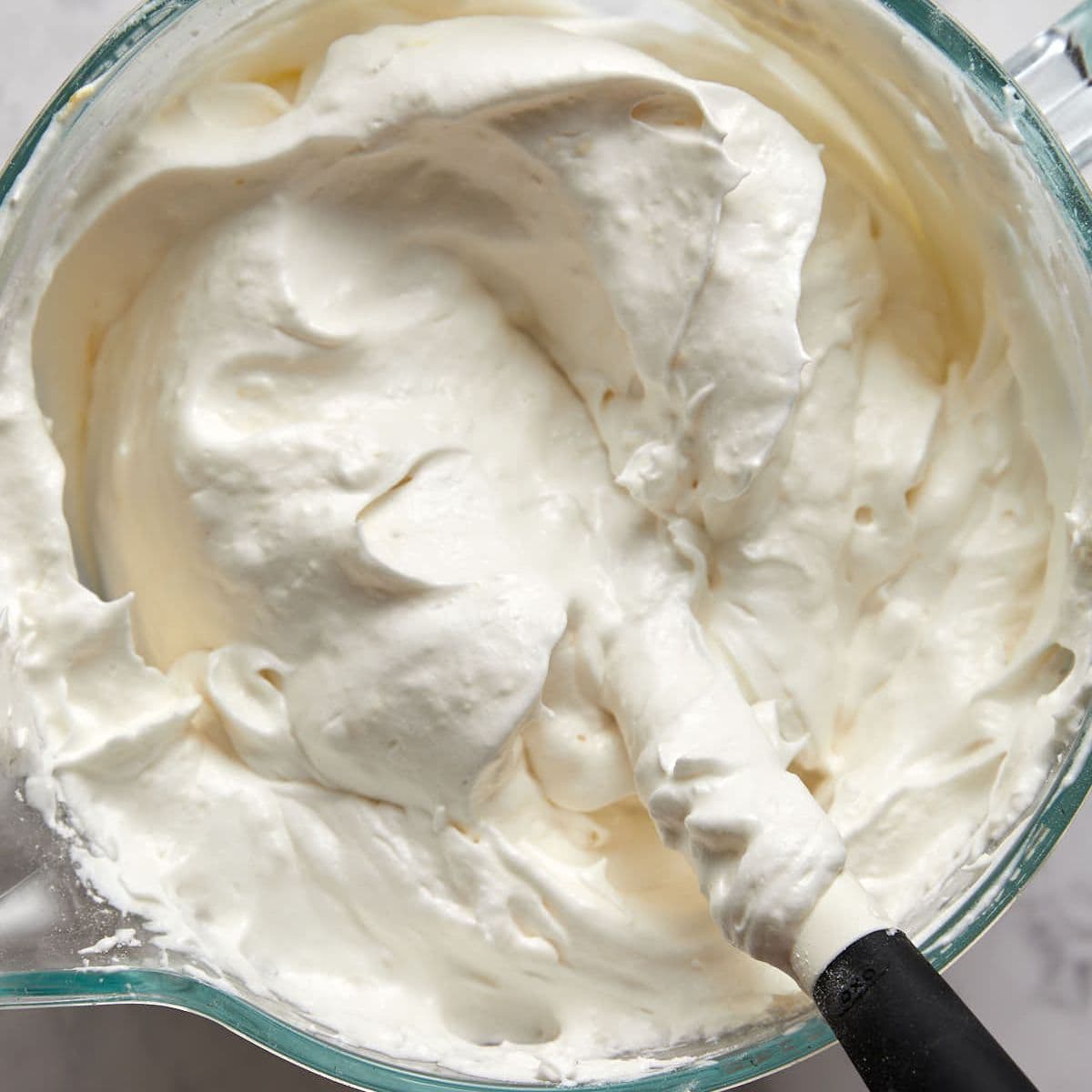 A glass mixing bowl filled with thick, fluffy whipped cream sits ready for your favorite angel food cake recipe. A spatula is partially submerged in the creamy mixture, showing its smooth and airy texture.