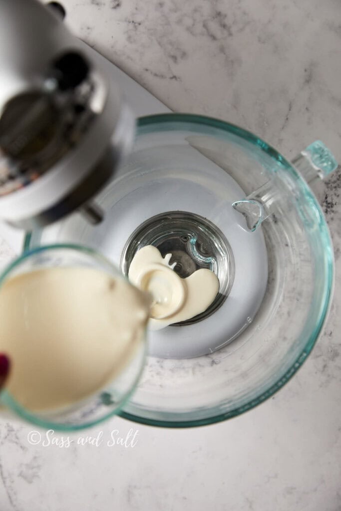 Overhead view of a stand mixer with a clear glass bowl on a marble counter. Someone is pouring a heavy cream into a mixer bowl.