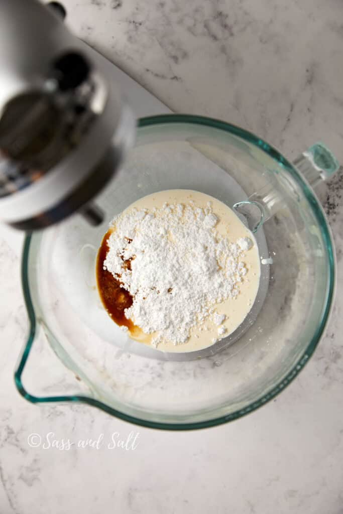 A stand mixer sits on a marble countertop, with a glass mixing bowl attached. Inside the bowl are ingredients, including cream, powdered sugar, and vanilla extract, ready to be mixed. The words "Sass and Salt" are visible near the bottom of the image.