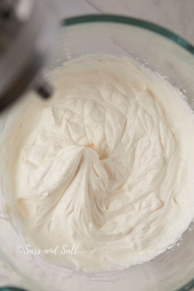 A glass bowl filled with fluffy whipped cream, captured from a top-angle view. The cream is smooth with soft peaks formed on the surface. A portion of a mixer is visible at the edge of the bowl.