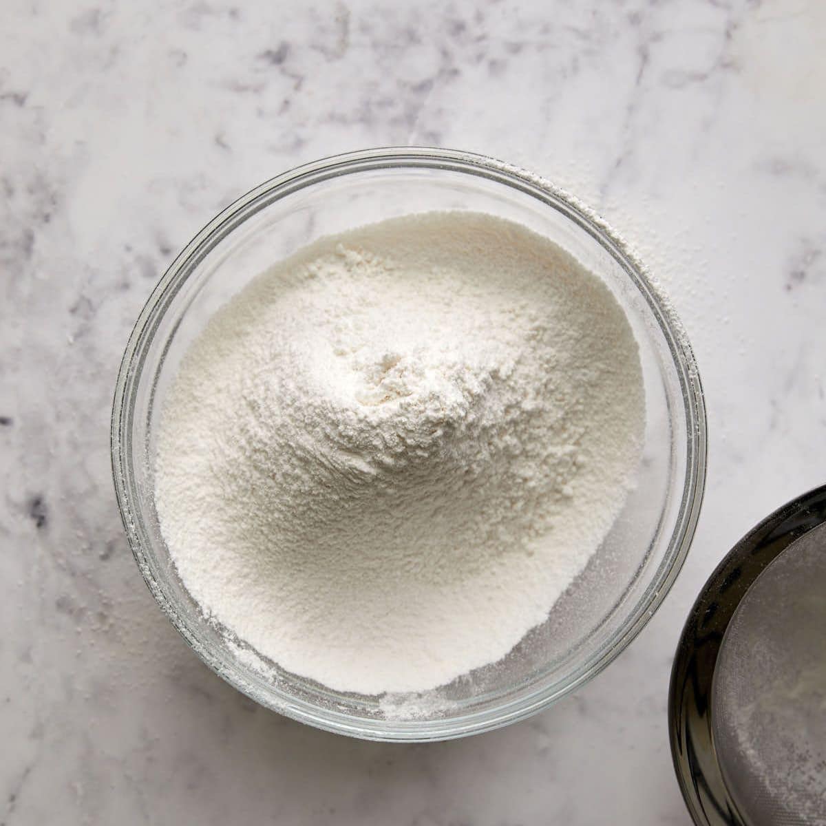 A clear glass bowl filled with white flour sits on a light marble surface. Some flour is scattered nearby.