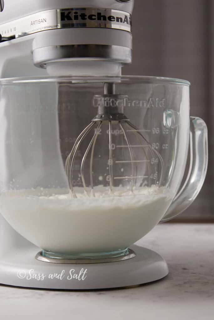 A white KitchenAid stand mixer, with a whisk attachment, mixing a heavy cream in a glass bowl on a marble countertop. The mixer branding is visible on the side and the bowl has measurement markings. The image is signed "Sass and Salt" at the bottom.