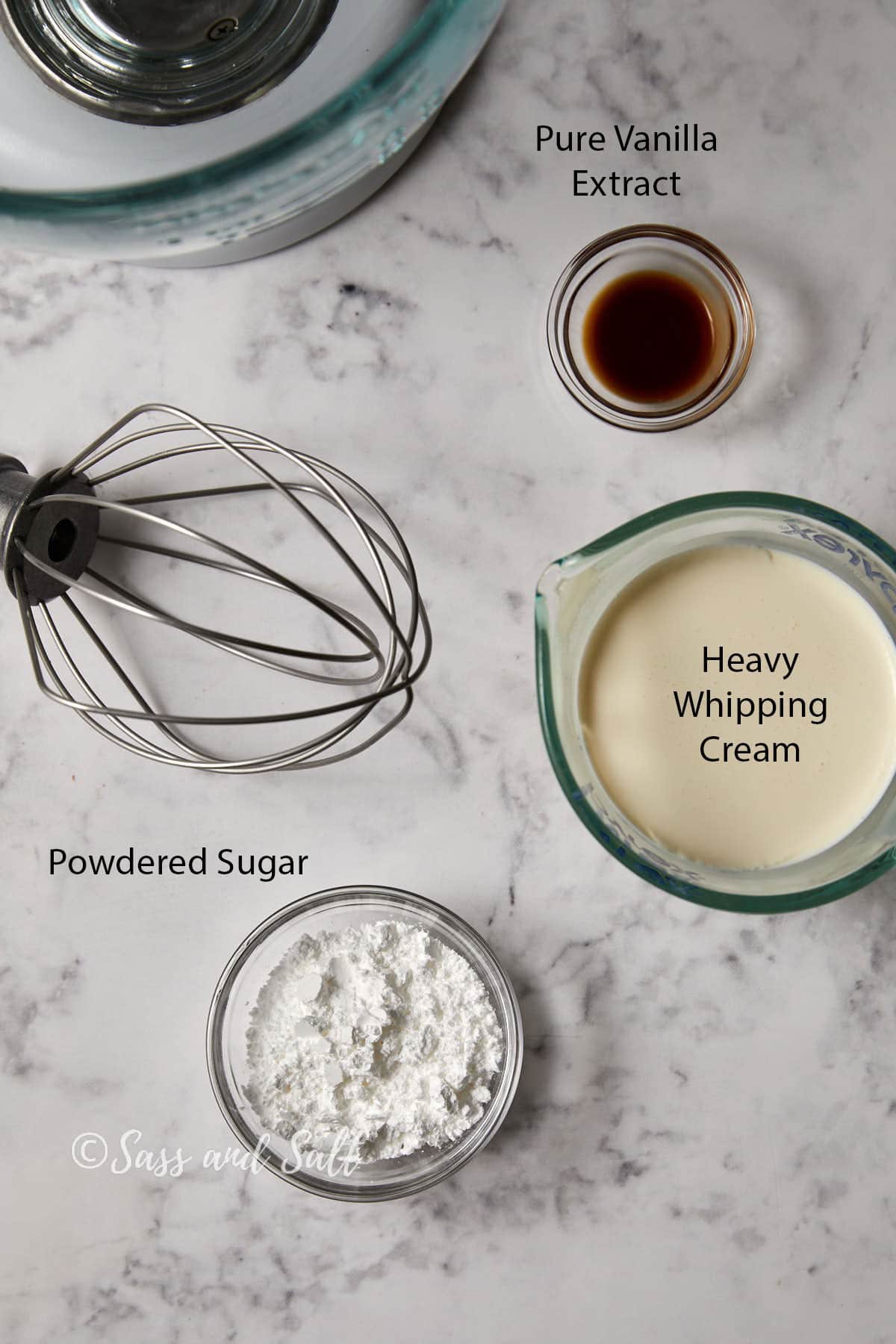 A marble surface with ingredients for making whipped cream: a small bowl of powdered sugar, a glass measuring cup of heavy whipping cream, a small bowl of pure vanilla extract, and a wire whisk attachment from a mixer. "Sass and Salt" is written at the bottom.