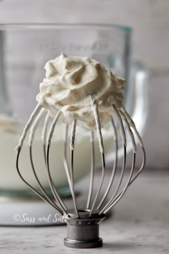 A whisk attachment, coated in whipped cream, is placed upright on a flat surface. In the blurry background, a glass mixing bowl filled with more whipped cream is visible. The image is bright and clean, suggesting a fresh kitchen scene.