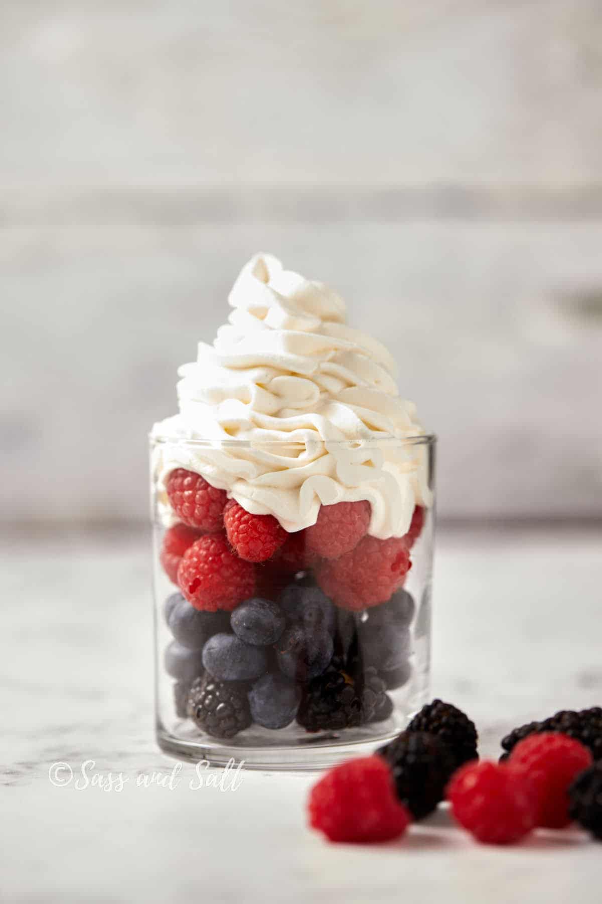 A glass cup filled with layers of blackberries, blueberries, and raspberries, topped with a generous swirl of whipped cream. The background is soft and neutral, emphasizing the vibrant colors of the berries. Loose raspberries and blackberries are scattered near the glass.