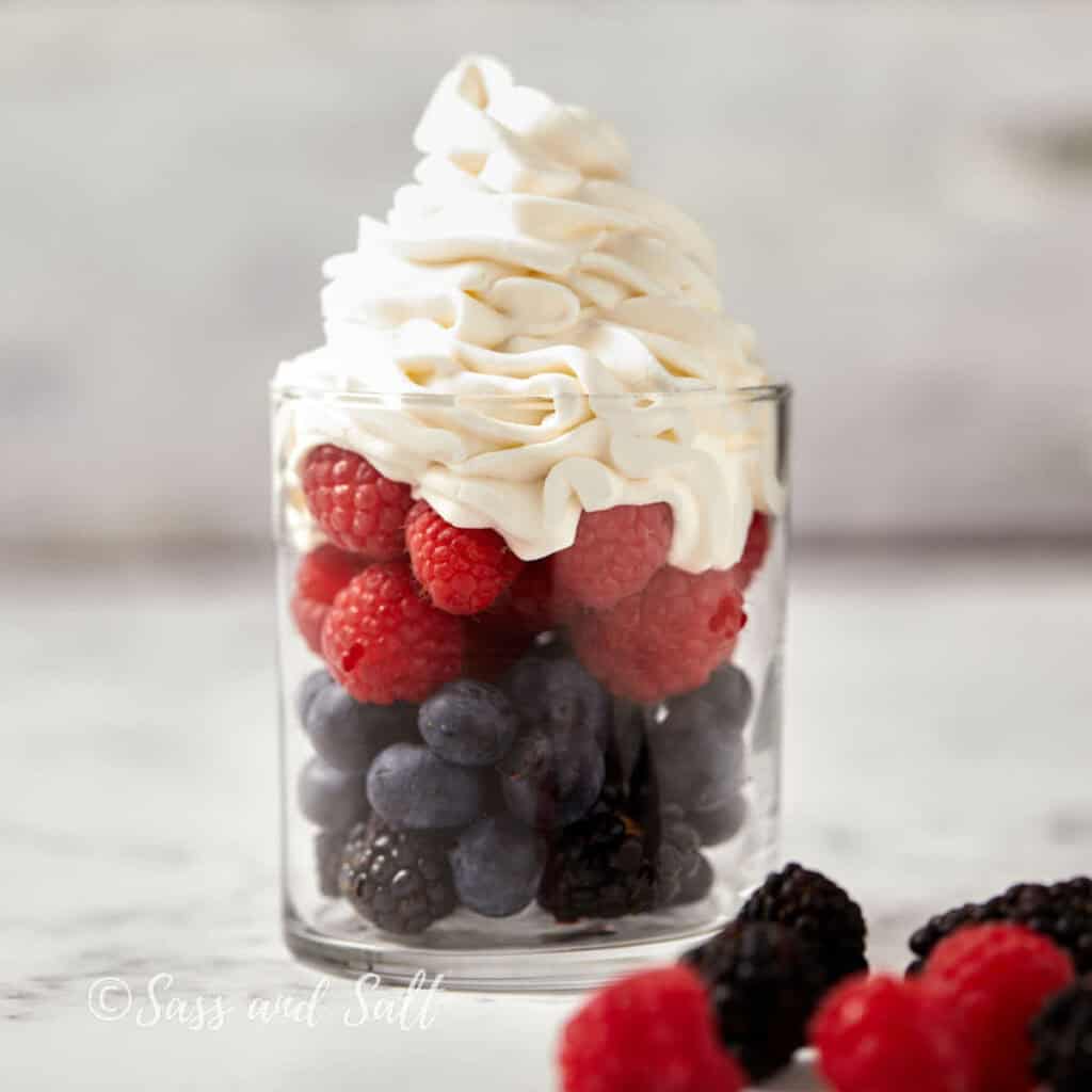 A clear glass filled with blueberries, blackberries, and raspberries, topped with a generous swirl of whipped cream, sits on a marble surface. Additional berries are scattered around the base of the glass. 