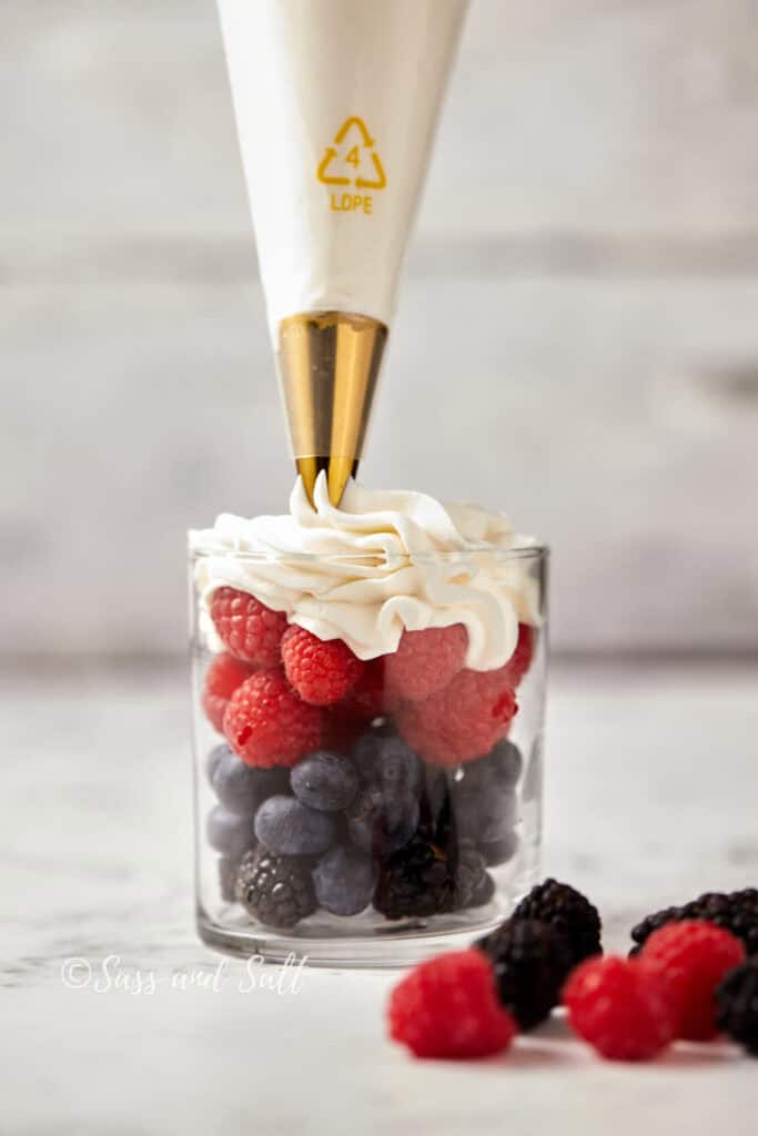 A glass filled with fresh blueberries, raspberries, and blackberries is topped with whipped cream from a piping bag. Additional berries are scattered beside the glass. The focus is on the whipped cream being added to the berries.