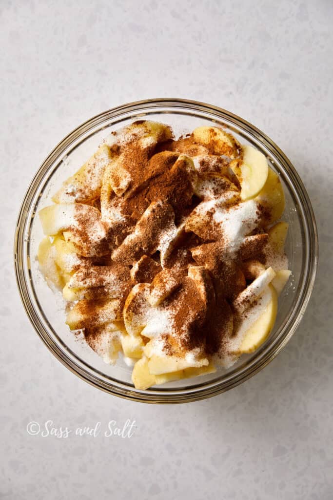 A glass bowl contains apple slices sprinkled with sugar, cinnamon and nutmg on top of the apple slices. The mixture is ready to be stirred. The background is a light-colored, speckled countertop.