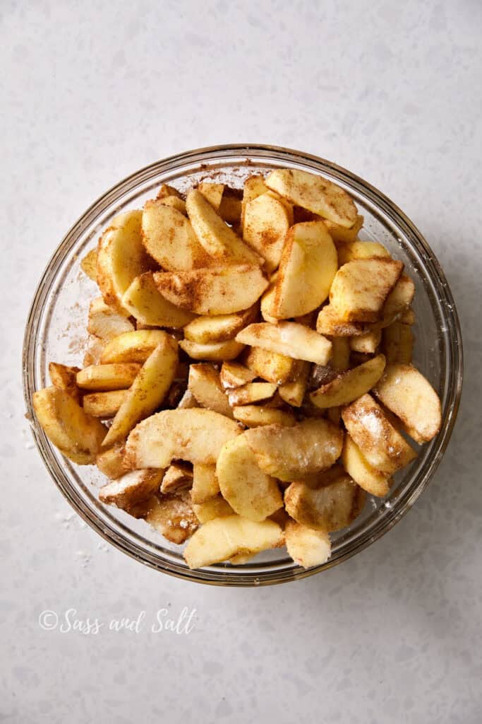 A top-down view of a glass bowl filled with sliced apples coated in cinnamon, nutmeg and sugar, placed on a white speckled surface.