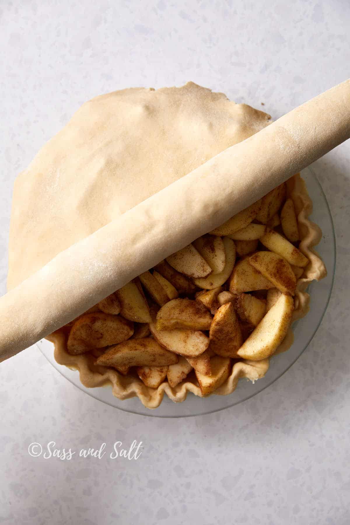 A partially covered apple pie with cinnamon-spiced apple slices is being prepared. A rolling pin with dough is positioned at the top, ready to cover the pie completely. The background is a speckled white surface. The text "Sass and Salt" is visible in the corner.