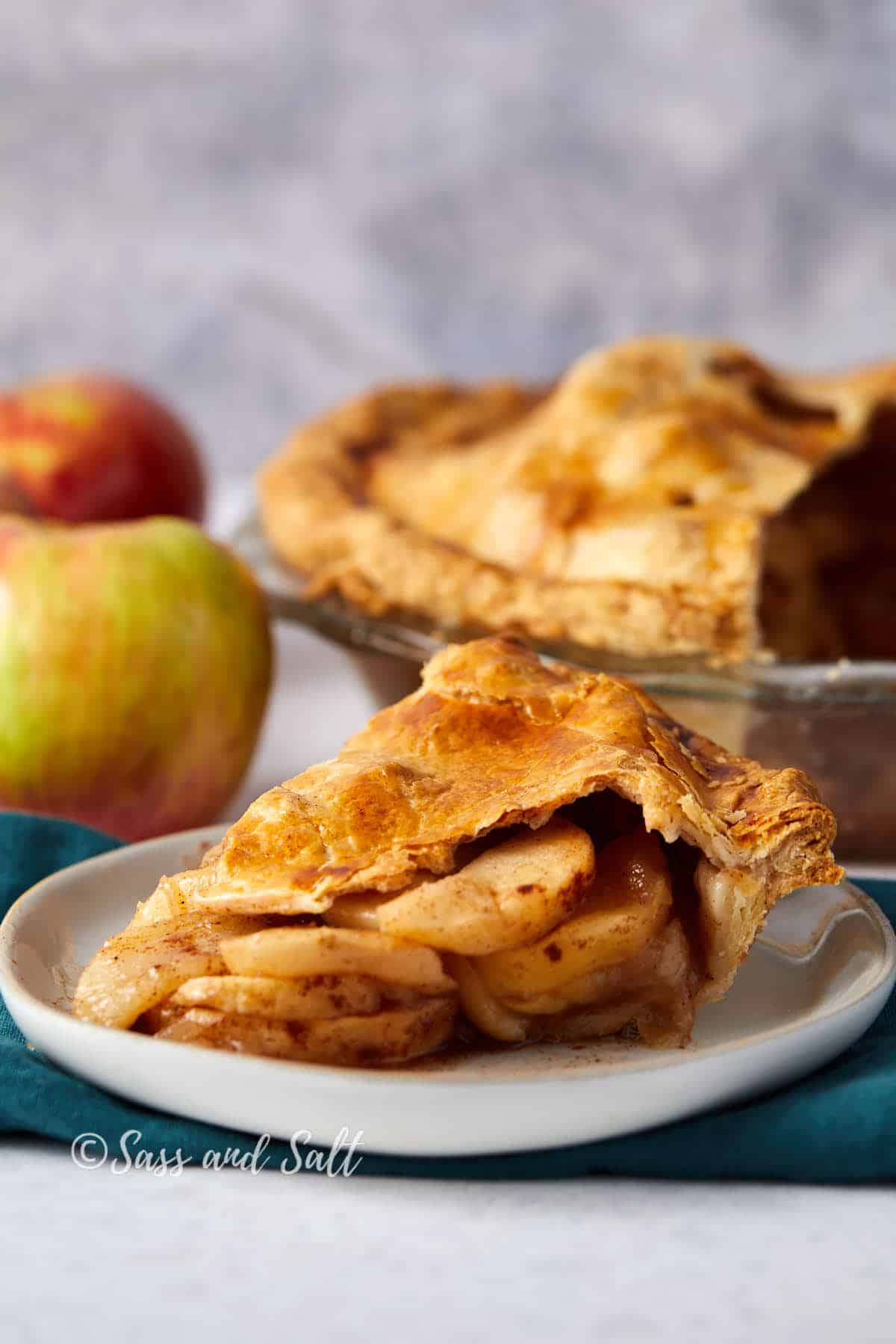 A slice of apple pie with a golden-brown crust is served on a white plate. The pie is filled with tender, spiced apple slices. The whole pie, partially sliced, is visible in the background along with a few fresh apples. The image is credited to "Sass and Salt.