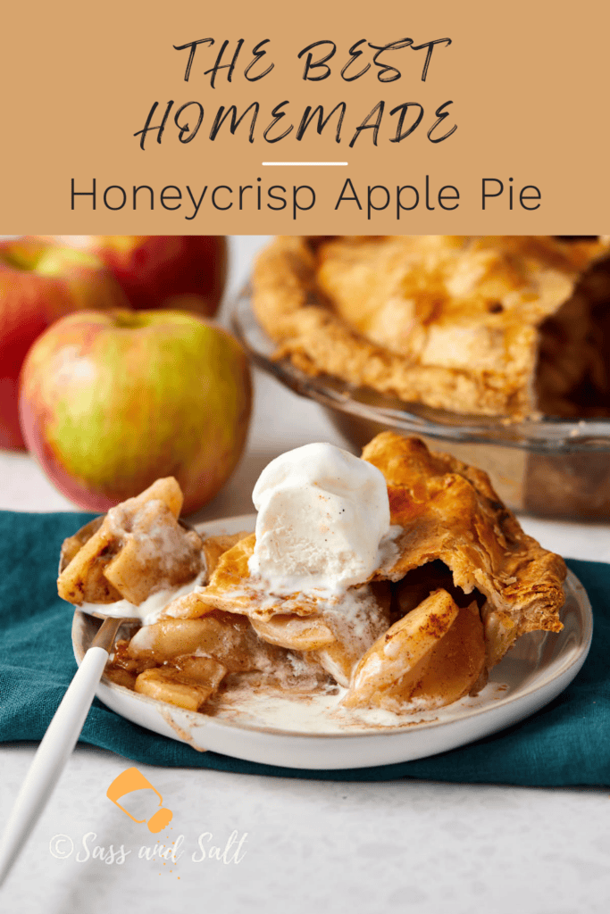 A slice of homemade Honeycrisp apple pie with a scoop of vanilla ice cream melting on top, served on a white plate. In the background, there are whole apples and the rest of the pie. Text overlay reads, "The Best Homemade Honeycrisp Apple Pie.