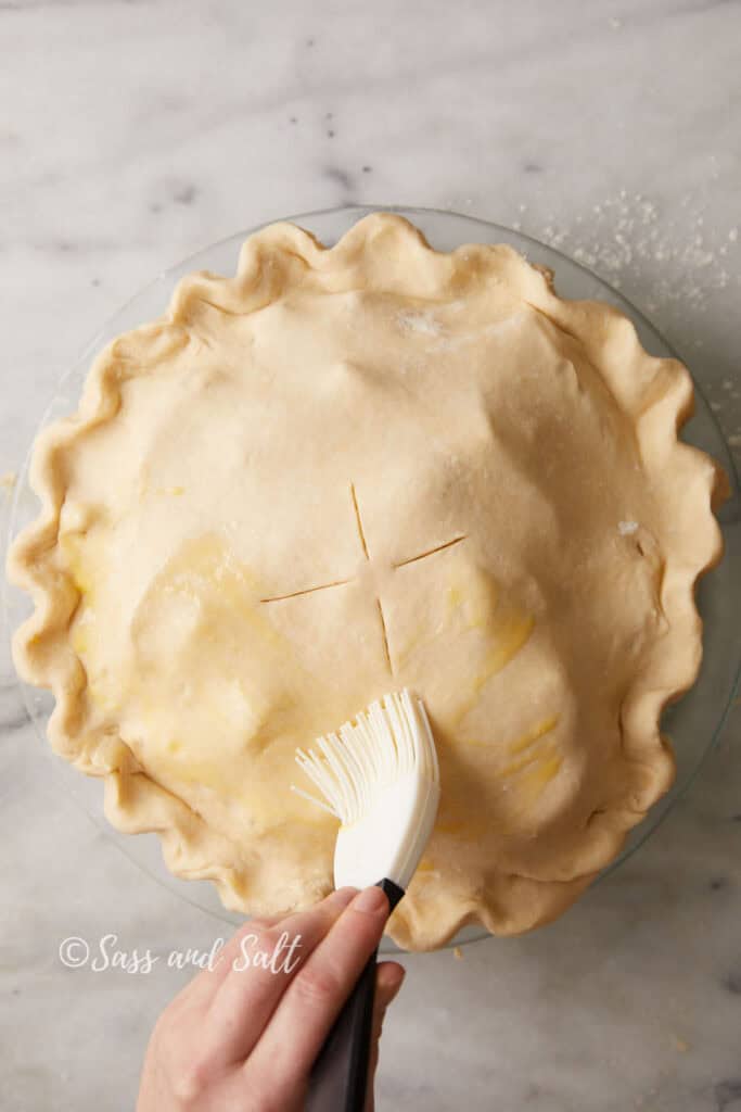 A person's hand is brushing egg wash onto the crimped edges of an uncooked pie crust with slits on top. The pie is placed on a marble surface.