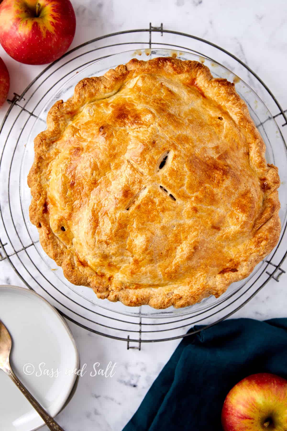 A golden-brown pie sits on a cooling rack atop a marble surface. Surrounding the pie are whole red apples, a dark blue cloth, and a white plate with a fork. The pie's crust looks flaky and inviting.