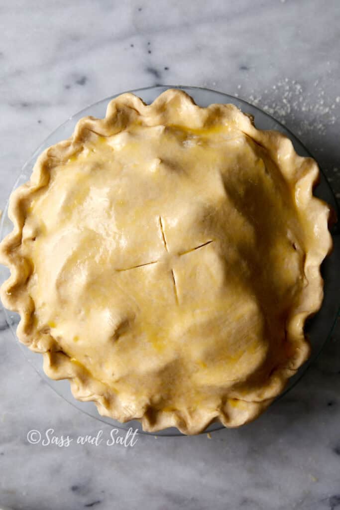 A pie with a golden, crimped crust and a cross-shaped vent on top sits on a marble surface. It appears freshly prepared and ready for baking. The image has a watermark that reads "Sass and Salt.